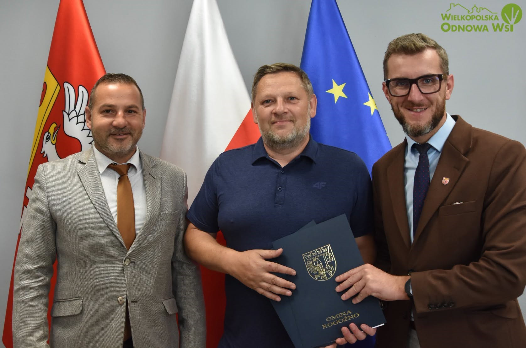 trzech mężczyzn stojących przodem do kamery, dw&oacute;ch trzyma teczkę z&nbsp;herbem Gminy Rogoźno, za nimi flagi Gminy Rogoźno, Polski i&nbsp;Unii Europejskiej