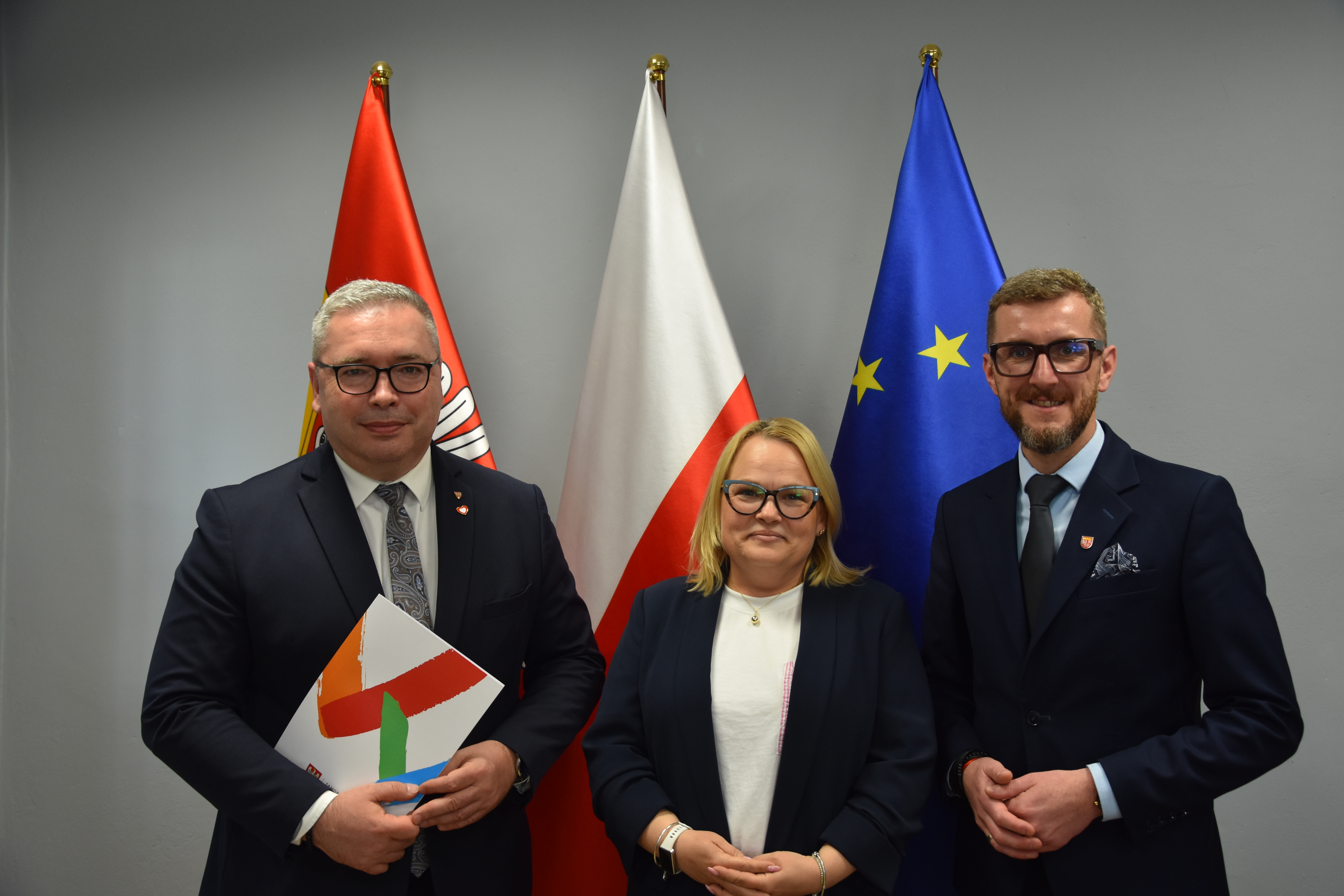 Troje ludzi w&nbsp;eleganckich strojach stoi na&nbsp;tle trzech flag - rogozińskiej, polskiej i&nbsp;europejskiej - po lewej i&nbsp;po prawej mężczyźni, po środku kobieta